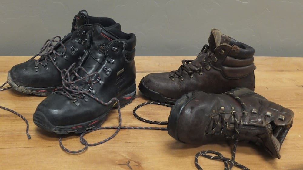 Two pairs of Zamberlan Boots, one black and one brown, sit on a wooden surface with their laces untied. Showing signs of decades of testing and use, these hiking boots tell a story of countless adventures.