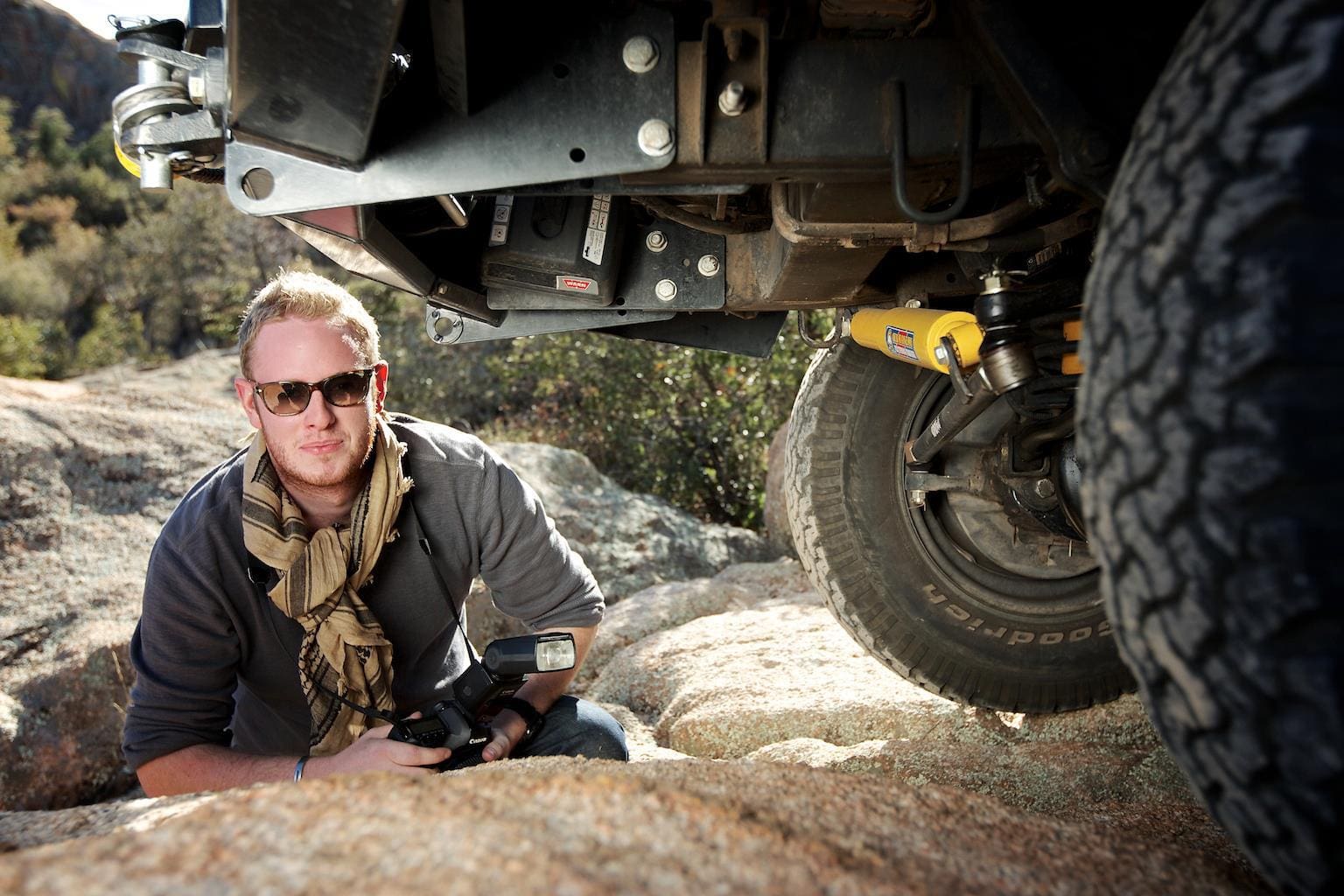 A person wearing sunglasses and a scarf kneels on rocks, holding a camera, next to an off-road vehicle with visible tire and suspension—getting electric with Rivian and Ford, as well as the powerful Hummer—in an outdoor setting framed by trees in the background.