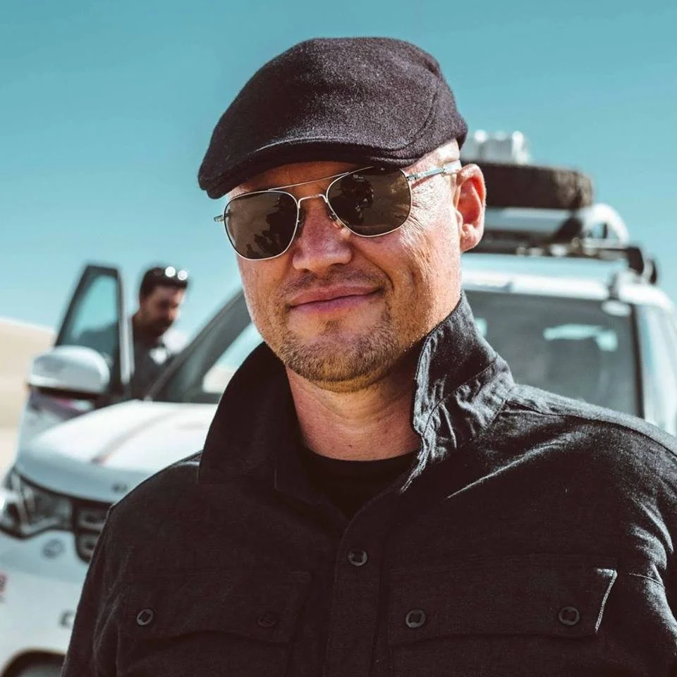 A man wearing sunglasses, a flat cap, and a dark jacket smiles in front of a white SUV parked in a sunny, outdoor setting—fully embracing the liberating leash of solo overlanding. Another person stands by the vehicle in the background, as if both are moments away from leaving the desk behind for adventure.