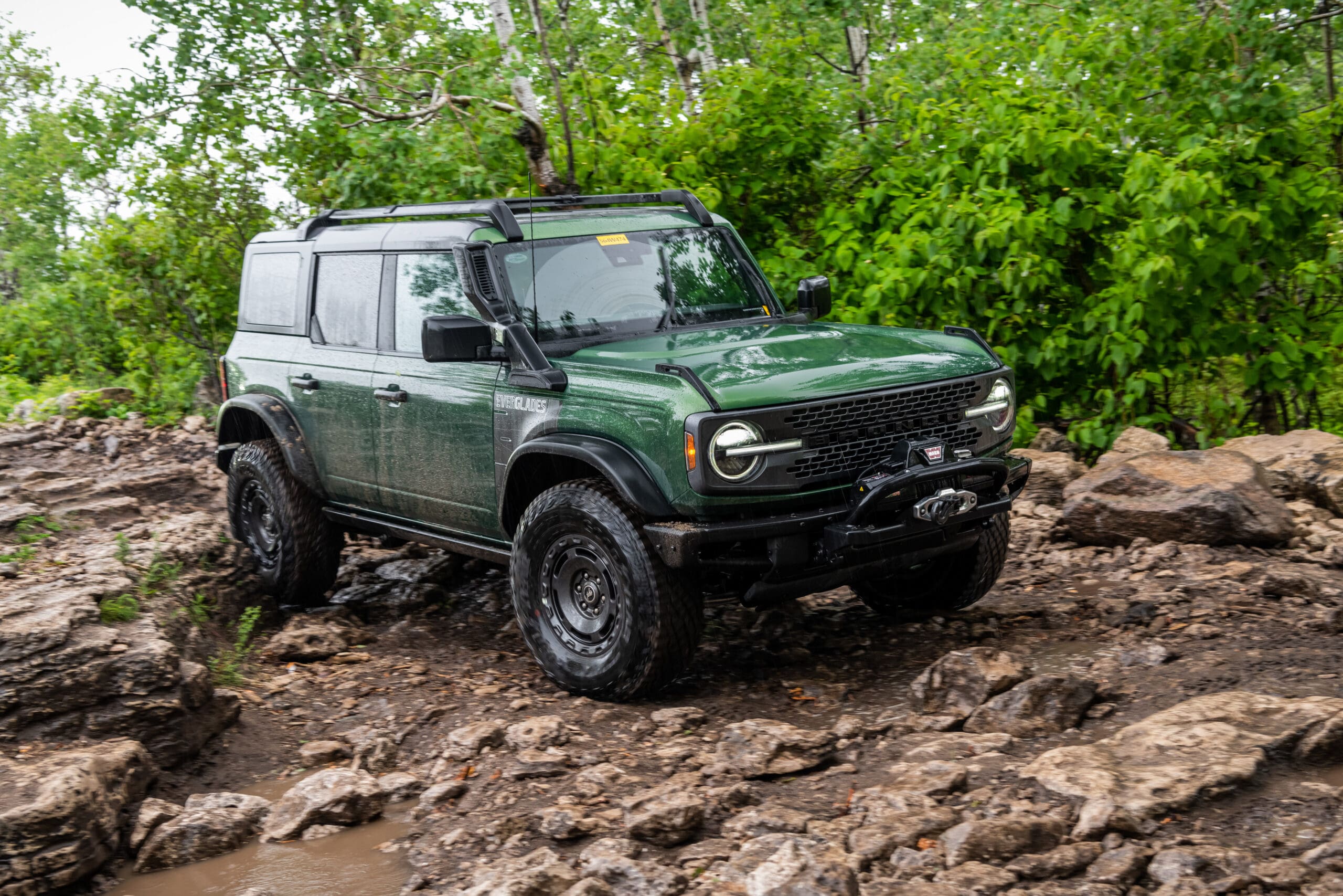 A green Ford Bronco SUV with off-road tires is tackling a rocky, muddy trail surrounded by dense green trees and foliage, showcasing the rugged capabilities of the new Bronco models.