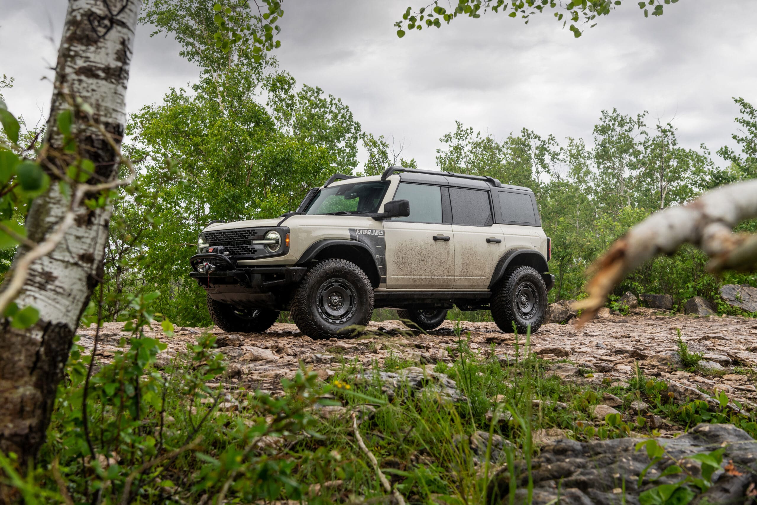 A white Ford Bronco with mud splatters is parked on a rocky, dirt path surrounded by green trees and plants under a cloudy sky, showcasing the rugged capability of Ford SUVs.