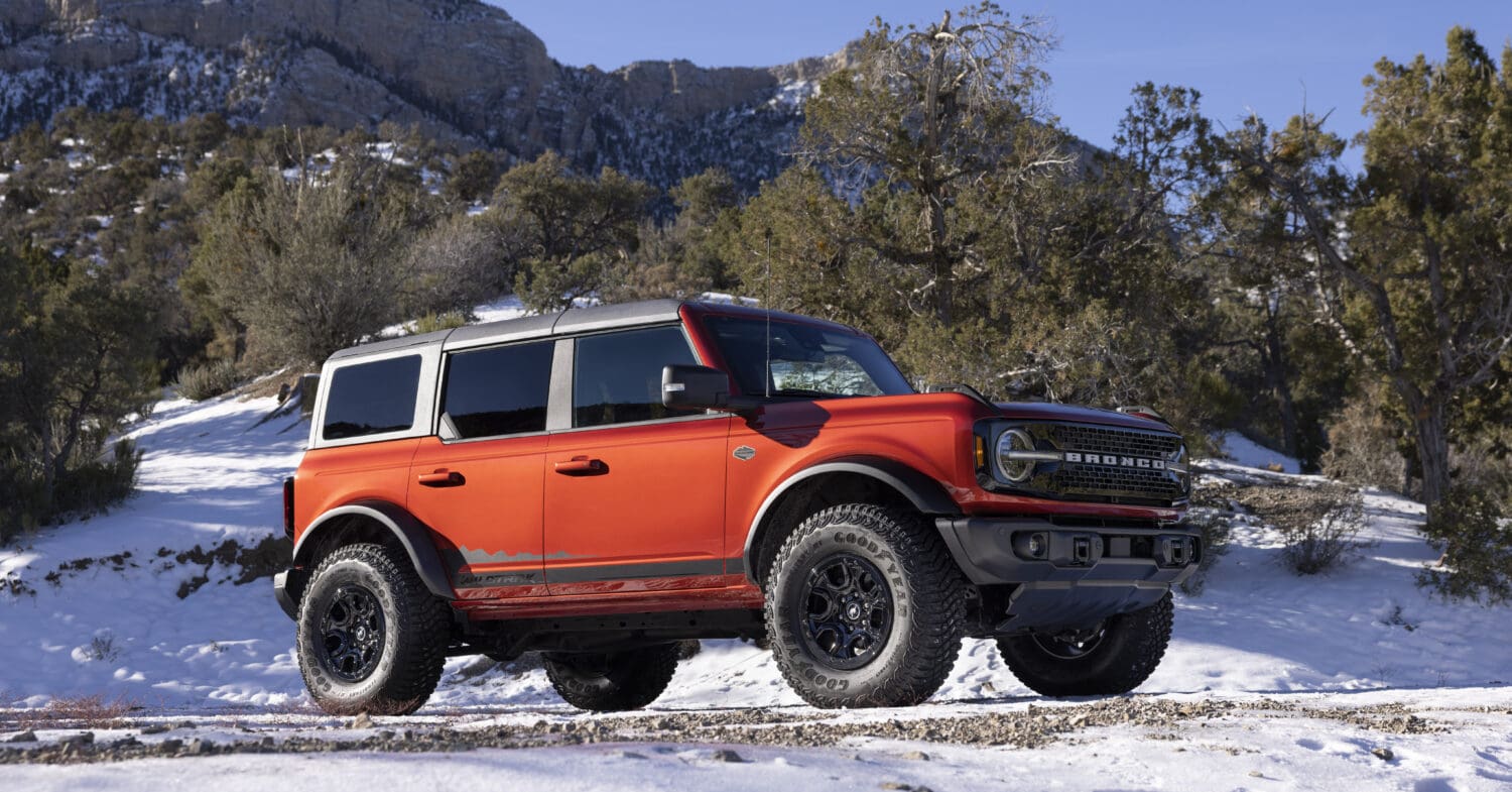 A bright orange Ford Bronco SUV is parked on a snowy dirt road with trees and rocky mountains in the background on a clear, sunny day, showcasing the adventurous spirit of Bronco models.