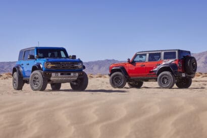Two Ford Bronco models, one blue and one red, parked on sandy desert terrain with tire tracks, mountains in the distance, and a clear blue sky above.