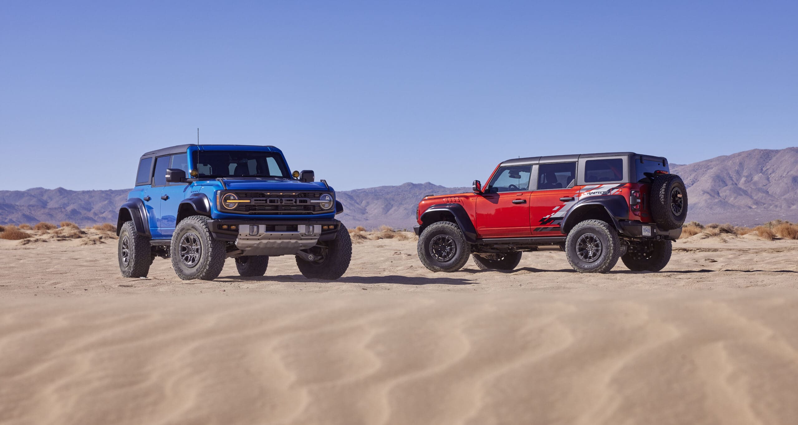 Two Ford Bronco models, one blue and one red, parked on sandy desert terrain with tire tracks, mountains in the distance, and a clear blue sky above.