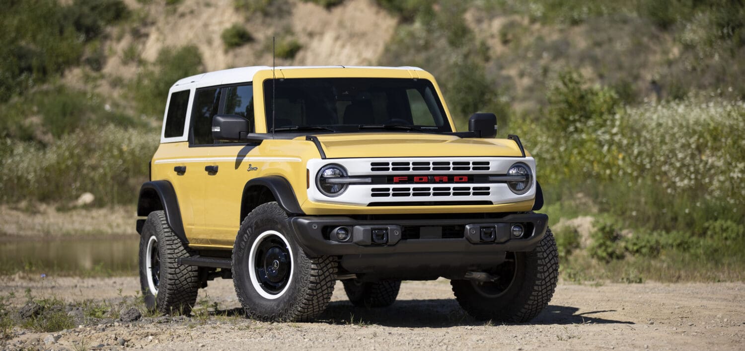 A yellow Ford Bronco SUV with a white roof is parked on a dirt path outdoors, surrounded by greenery and a rocky hillside in the background, showcasing the rugged appeal of new Bronco models.