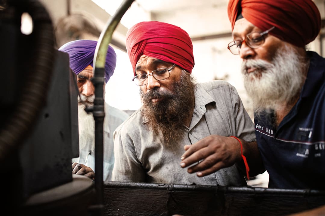 Three elderly men wearing colorful turbans and glasses closely examine machinery in an industrial setting, appearing focused and engaged—like seasoned engineers from Ladakh or travelers aboard the Wonder Bus through Kashmir.