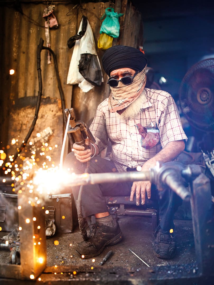 A man wearing dark glasses, a turban, and a scarf over his face welds metal, creating bright sparks in a Ladakh workshop surrounded by tools and industrial equipment.