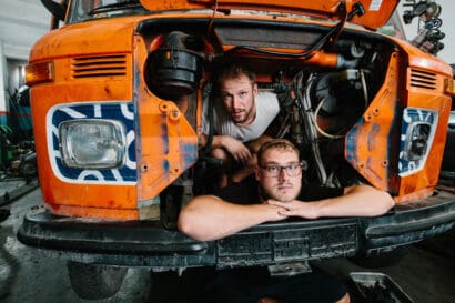 Two men pose inside the open hood of an old, orange Wonder Bus in a garage. One crouches in the engine bay while the other leans on the front bumper, both looking at the camera, capturing a quirky moment before their Ladakh adventure.