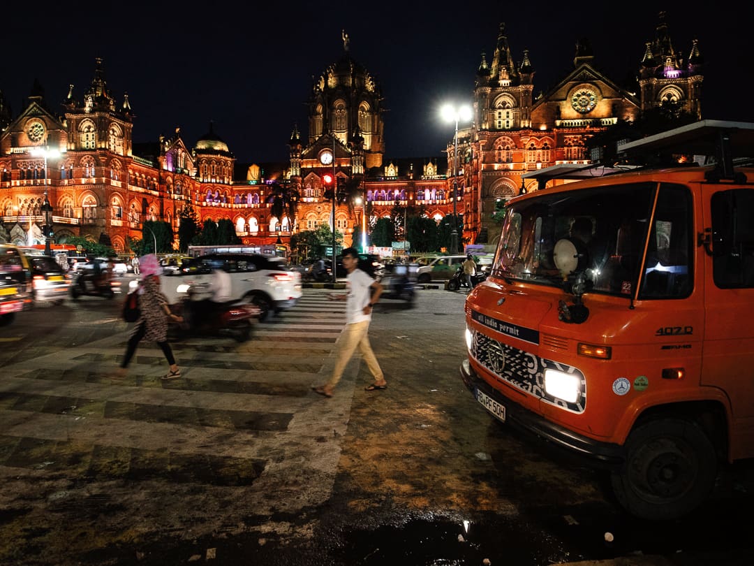 People cross a busy street at night in front of the brightly illuminated Chhatrapati Shivaji Maharaj Terminus in Mumbai, India, with traffic, an orange truck, and a Wonder Bus offering trips to Ladakh and Kashmir visible in the foreground.