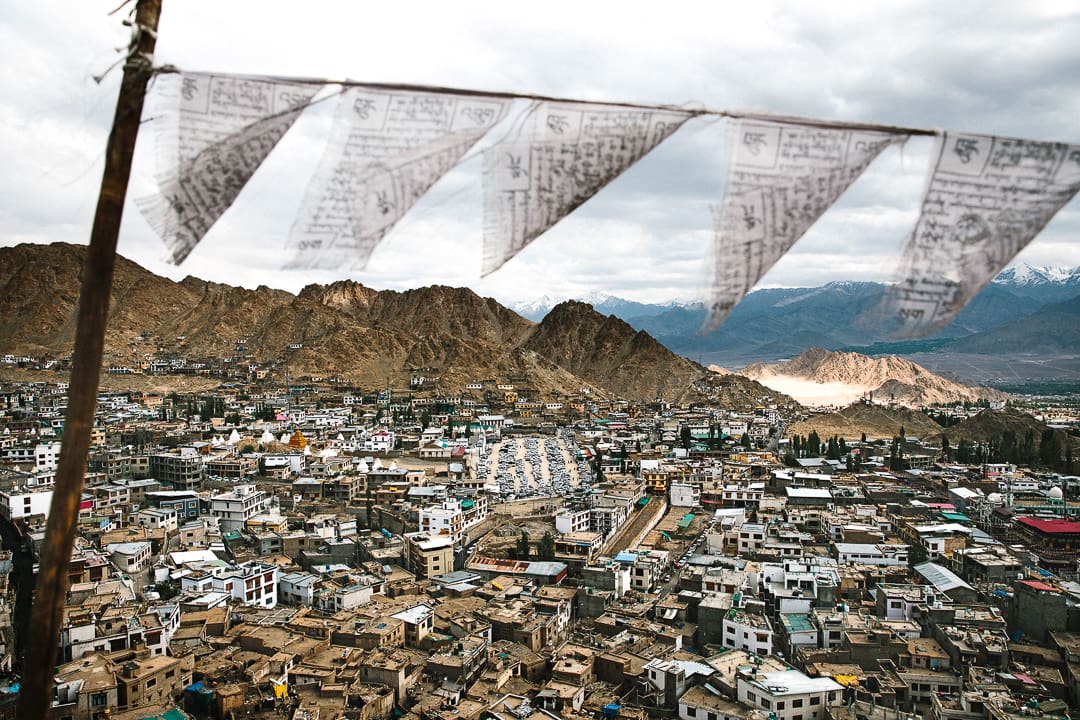 Prayer flags flutter above a densely packed Ladakh mountain town, with brown, rugged hills in the background and snow-capped peaks of Kashmir visible in the distance under a cloudy sky.
