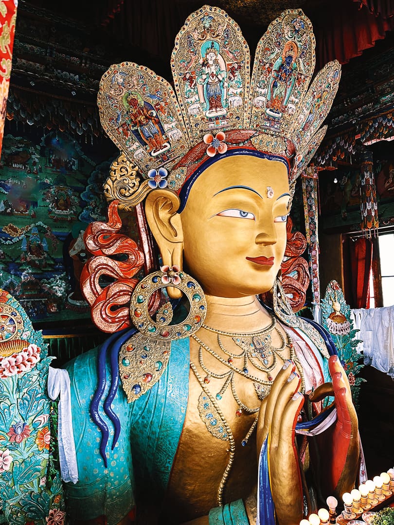 A large, ornate statue of Maitreya Buddha with a golden face, intricate crown, and colorful jewelry sits in a richly decorated Ladakh temple interior, capturing the region's unique spiritual heritage.