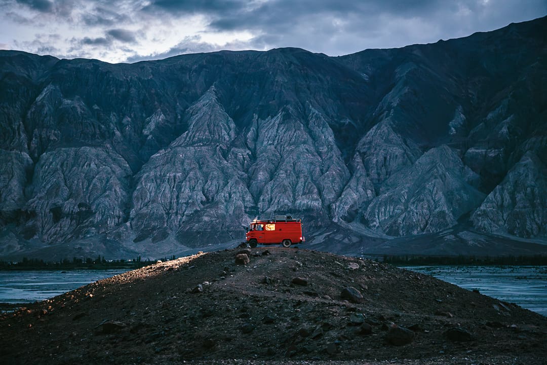 A red Wonder Bus is parked on a small hill at dusk, illuminated by soft light, with dramatic rocky mountains and a cloudy Kashmir sky in the background.