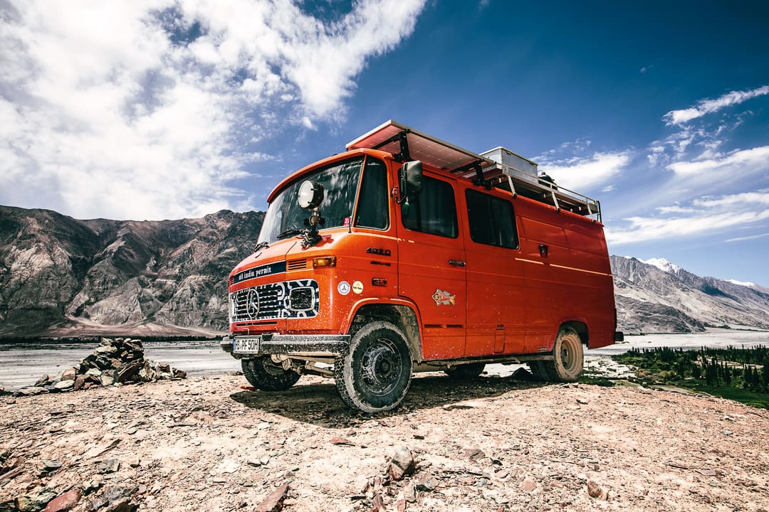 A bright orange Wonder Bus camper van parked on rocky terrain with a mountainous Ladakh landscape and blue sky in the background. The van is dusty and equipped with a roof rack and solar panels.