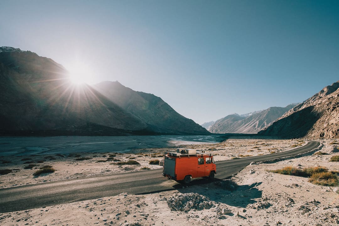 A bright orange Wonder Bus travels on a winding road through Ladakh's rocky, mountainous landscape, with the sun rising over distant peaks and casting long shadows across the valley.