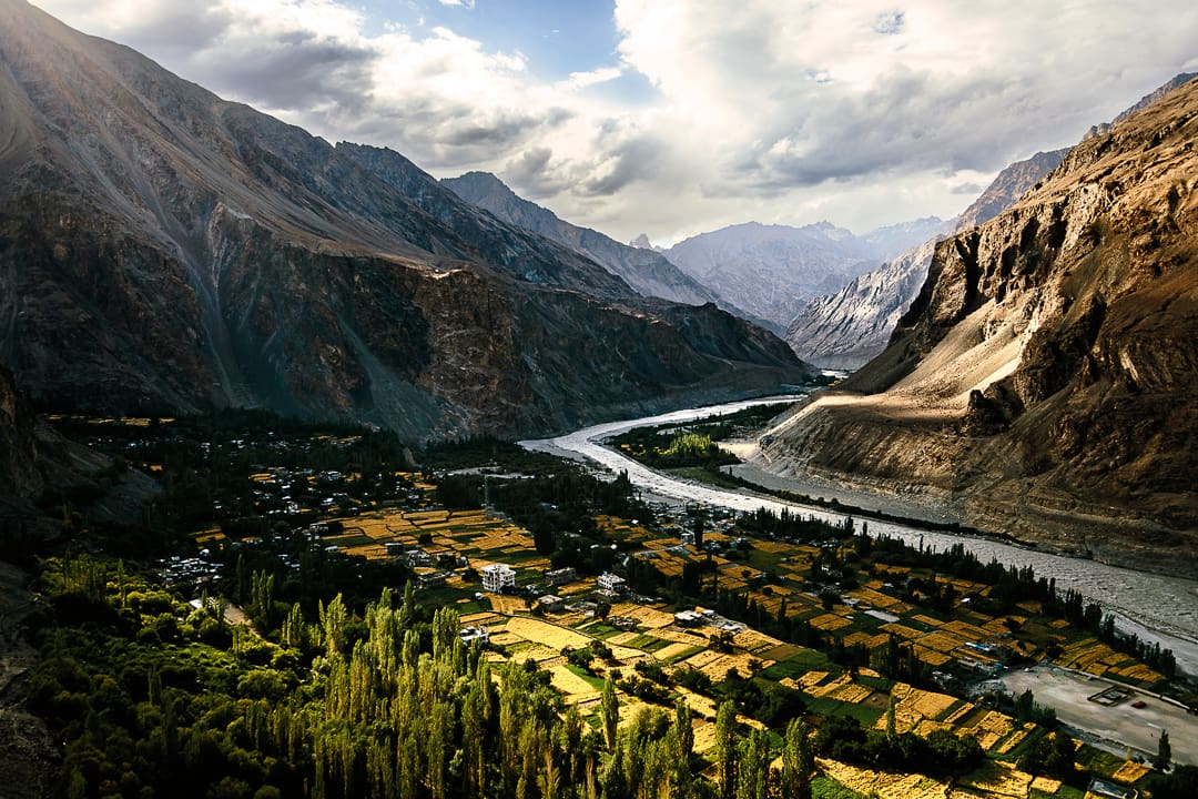 A river winds through a lush, green valley with yellow fields and trees in Kashmir, surrounded by tall, rocky mountains under a partly cloudy sky.