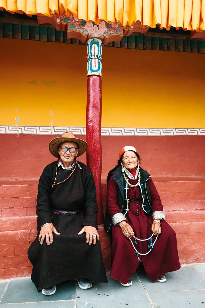 An elderly man and woman in traditional Tibetan clothing sit side by side on a red bench against a colorful wall in Ladakh, separated by a red pillar. Both wear hats and layered jewelry, gazing calmly at the camera.
