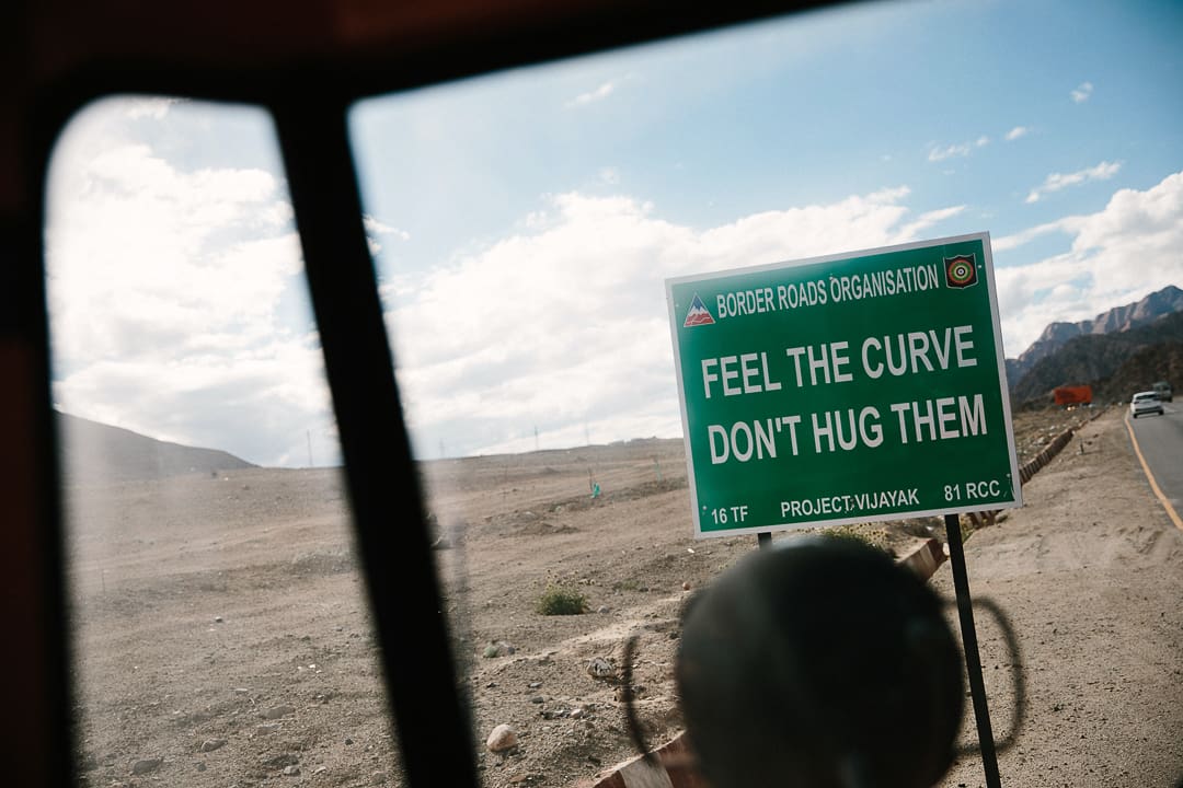 A green roadside sign reads FEEL THE CURVE, DONT HUG THEM beside a desert road in Ladakh under a partly cloudy sky, viewed through a Wonder Bus window.