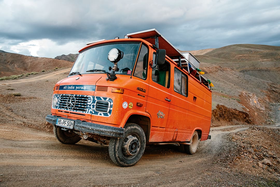An orange Wonder Bus with roof racks and solar panels drives on a rough dirt road through the rugged mountains of Ladakh under a cloudy sky.
