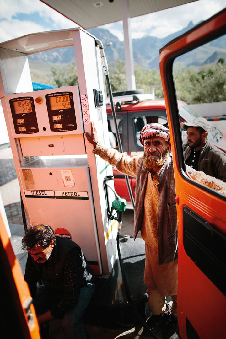 A man in traditional clothing smiles while holding a petrol pump beside an orange Wonder Bus at a gas station; another man kneels by the vehicle, with breathtaking mountains of Ladakh visible in the background.