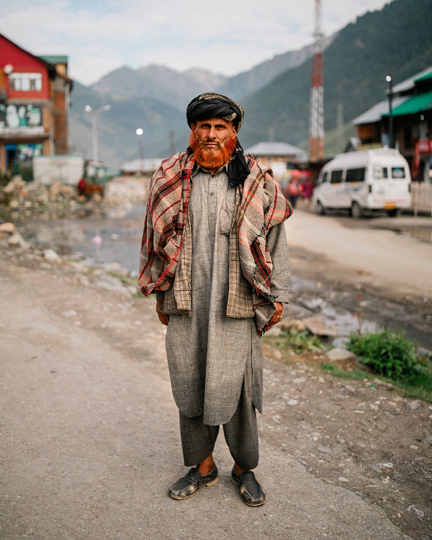 An elderly man with a bright orange beard, wearing traditional gray clothing, a checked shawl, and a black cap, stands on a street in Kashmir with mountains, buildings, vehicles, and the Wonder Bus in the background.