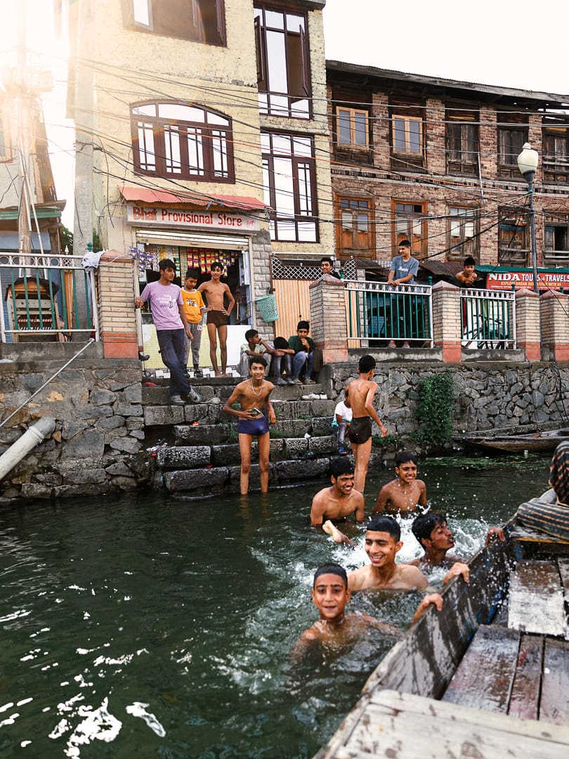 A group of boys swim and play in a canal near stone steps in Kashmir, while others watch from the steps and a bridge. Older buildings and a “Bhat Provisional Store” storefront are visible in the background.