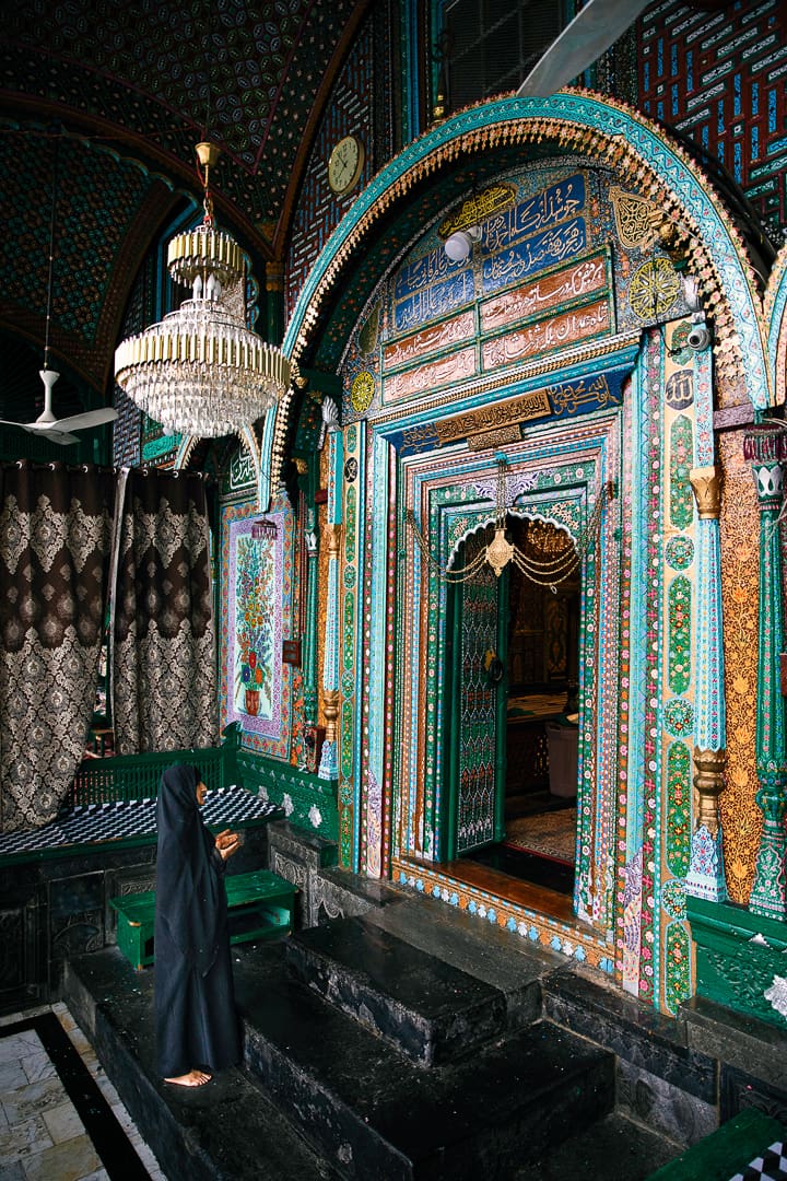 A person in a black robe stands in prayer before a richly decorated, colorful doorway inside a mosque in Kashmir, surrounded by intricate patterns, calligraphy, and a chandelier above.