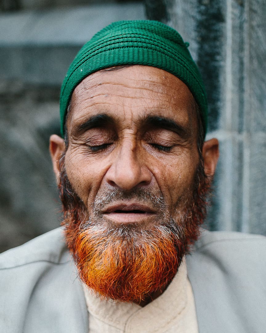 An older man with a dyed orange beard and a green knit cap sits with his eyes closed, wearing a pale jacket and beige shirt, in front of a stone background—capturing the tranquil spirit of Ladakh.