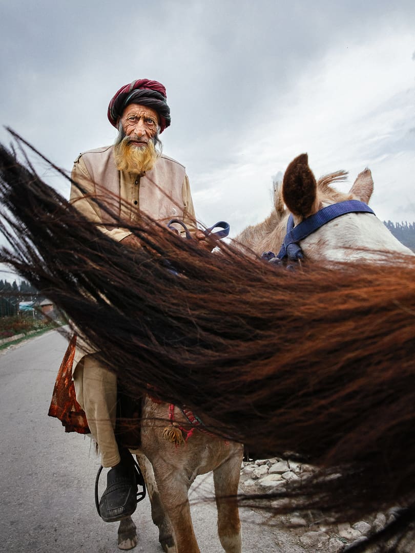 An elderly man with a turban and long white beard rides a horse on a rural Ladakh road, partially obscured by the horse’s flowing tail in the foreground under a cloudy sky.