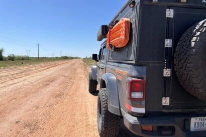A gray off-road vehicle with Texas plates is parked on a dirt road beneath a clear blue sky, its recovery gear ready for adventure. The scene is reminiscent of classic Overland Routes through grassy fields and power lines, like those in Iberá Park, Northeast Argentina.