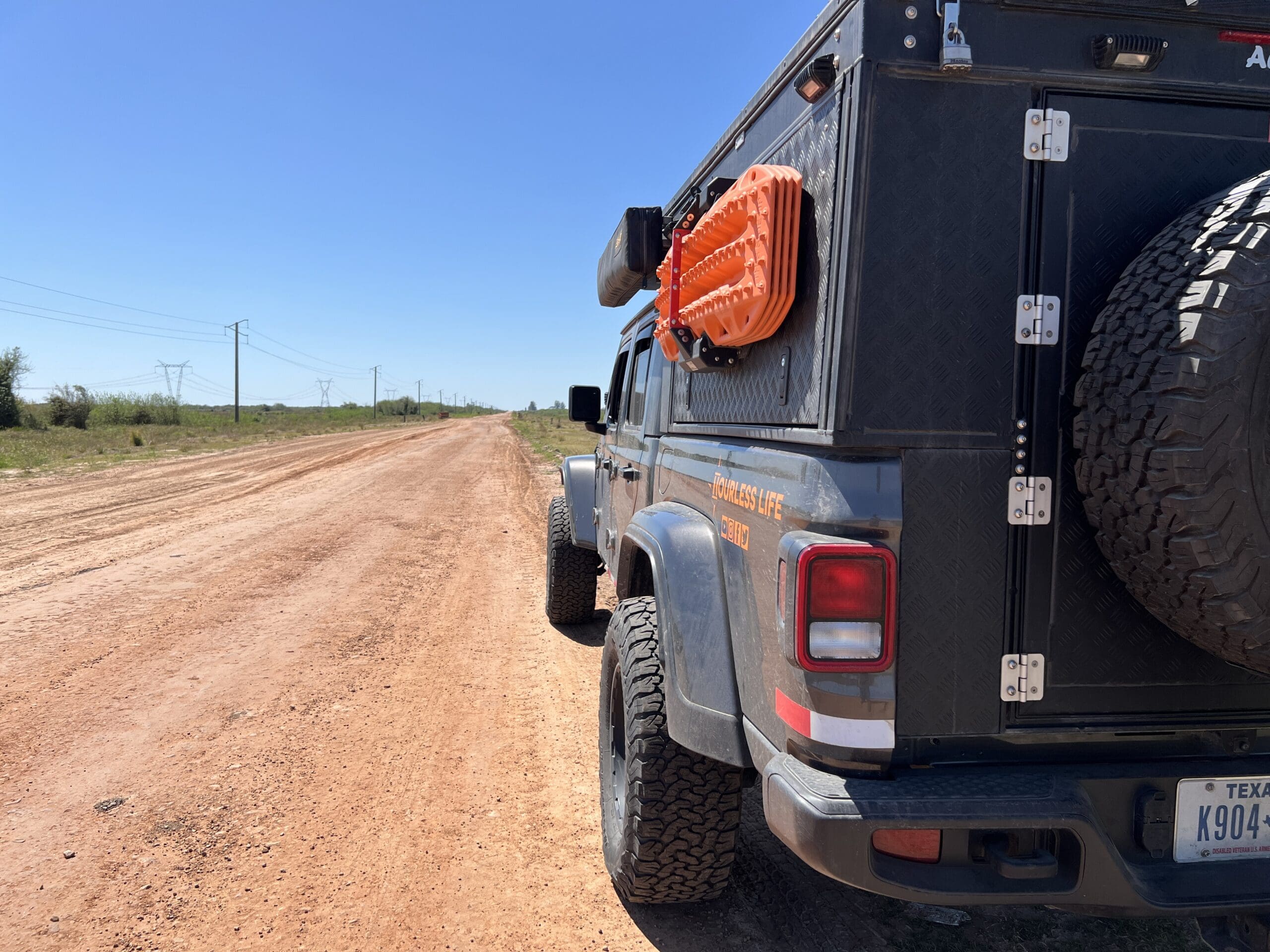 A gray off-road vehicle with Texas plates is parked on a dirt road beneath a clear blue sky, its recovery gear ready for adventure. The scene is reminiscent of classic Overland Routes through grassy fields and power lines, like those in Iberá Park, Northeast Argentina.