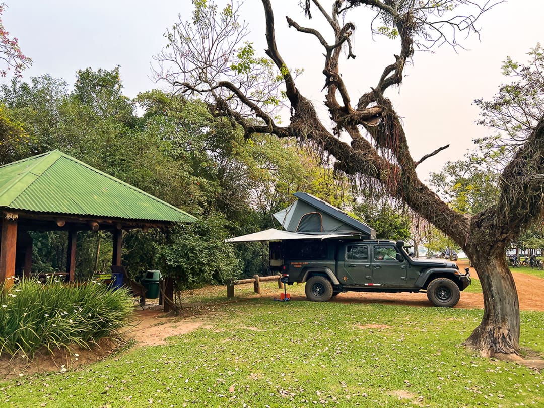 A green off-road vehicle with a rooftop tent is parked on grass near a rustic gazebo in Iberá Park, Northeast Argentina, surrounded by trees and greenery—an inviting stop along scenic overland routes on a clear day.