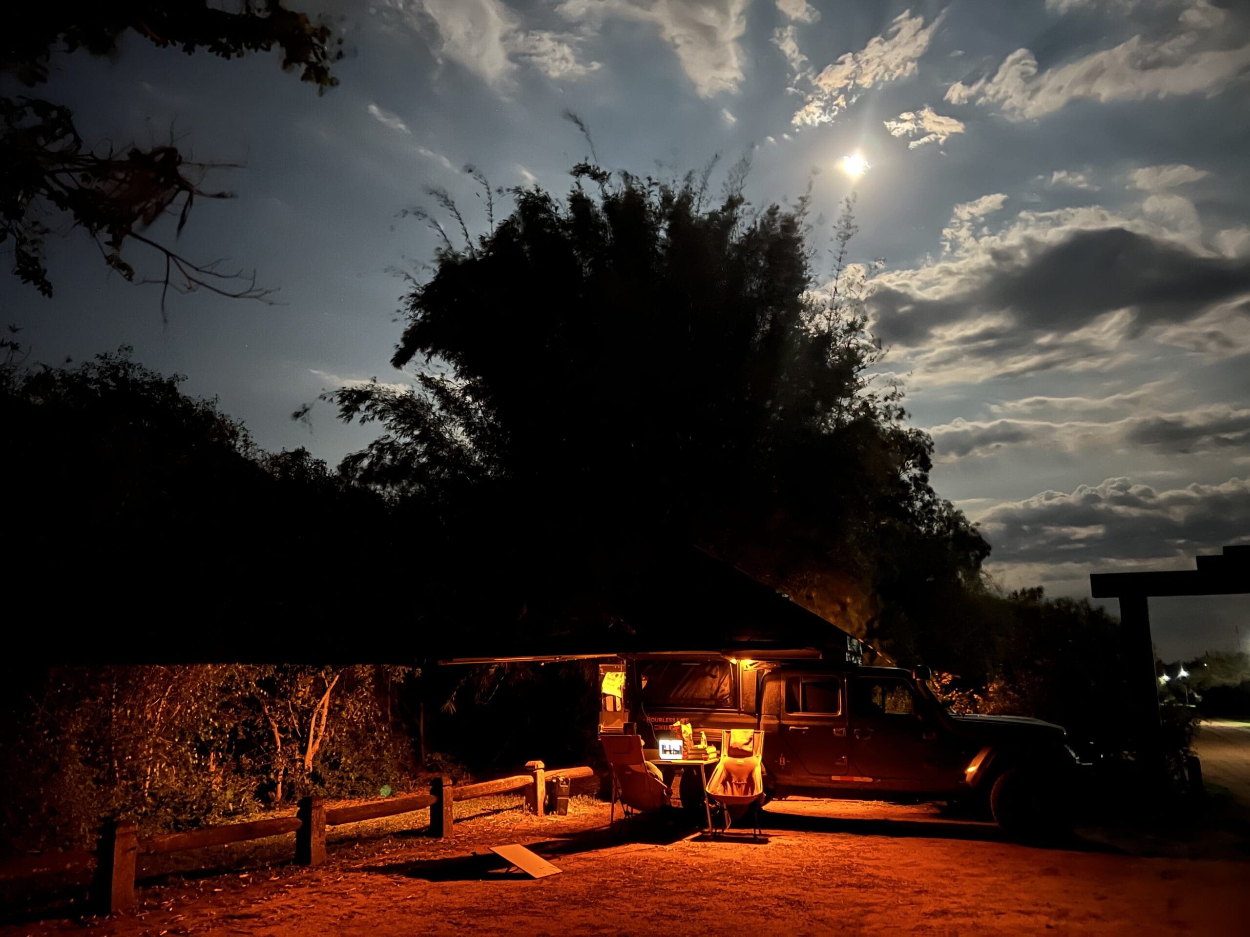 A Jeep with an awning is parked under trees at night along scenic Overland Routes. Warm orange lights illuminate the campsite, while the moon shines brightly behind clouds in the dark, partly cloudy sky of Northeast Argentina.