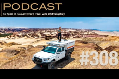 A woman stands on top of a white off-road camper truck in a remote, colorful desert landscape with rocky hills. Text reads, PODCAST: Six Years of Solo Adventure Travel with @hifromashley #308.