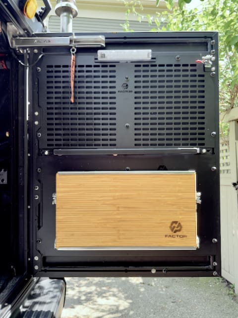 An open Toyota Tundra door reveals a black metal interior panel with ventilation holes and a mounted wooden fold-out table featuring the FACTOR logo, set outdoors by trees and a fence, showcasing the 2024 Alu-Cab Alu-Cabin setup.