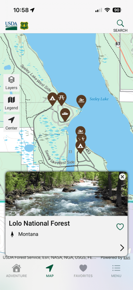 A smartphone screen displays a map of Seeley Lake in Lolo National Forest, Montana, with Forest Service icons for camping, hiking, fishing, and boating. At the bottom, a river photo is labeled Lolo National Forest.