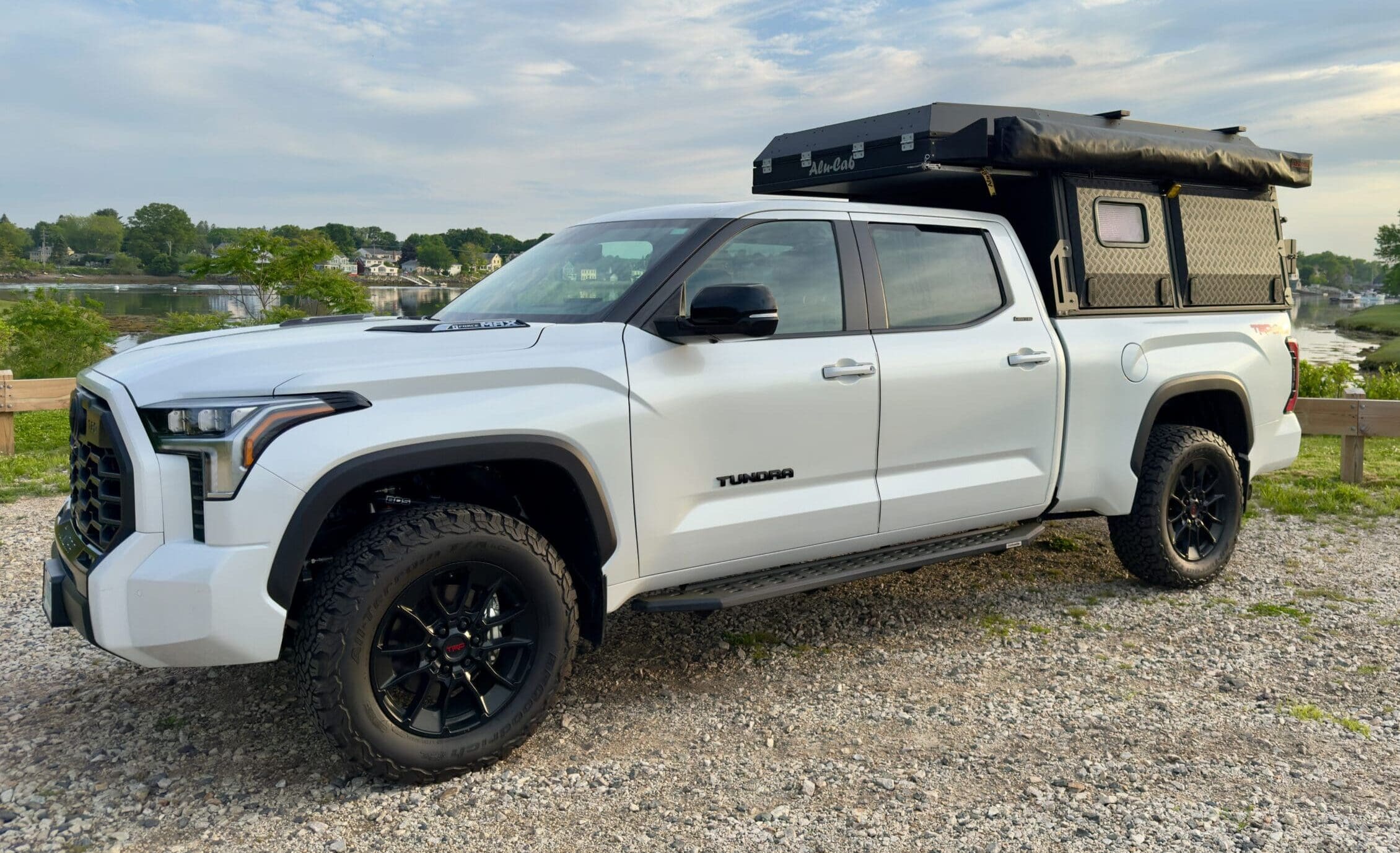 A white Toyota Tundra pickup truck with a black Alu-Cab camper shell is parked on gravel near a wooden fence. Trees, water, and houses are visible in the background under a partly cloudy sky.