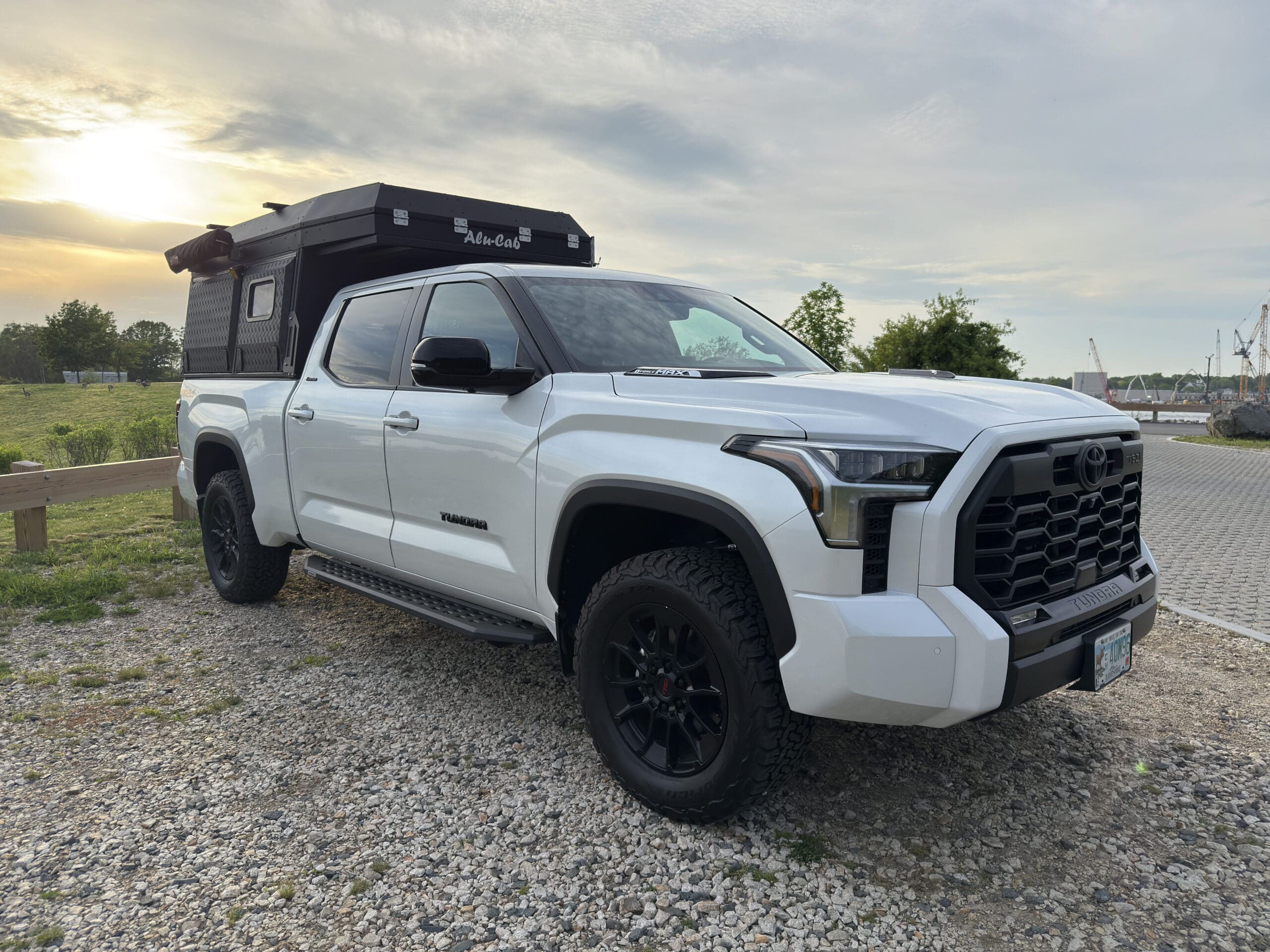 A white 2024 Toyota Tundra with black wheels is parked on a gravel lot at sunset, featuring a rugged Alu-Cab Alu-Cabin camper attached to its bed. Trees, grass, and a paved area can be seen in the background.
