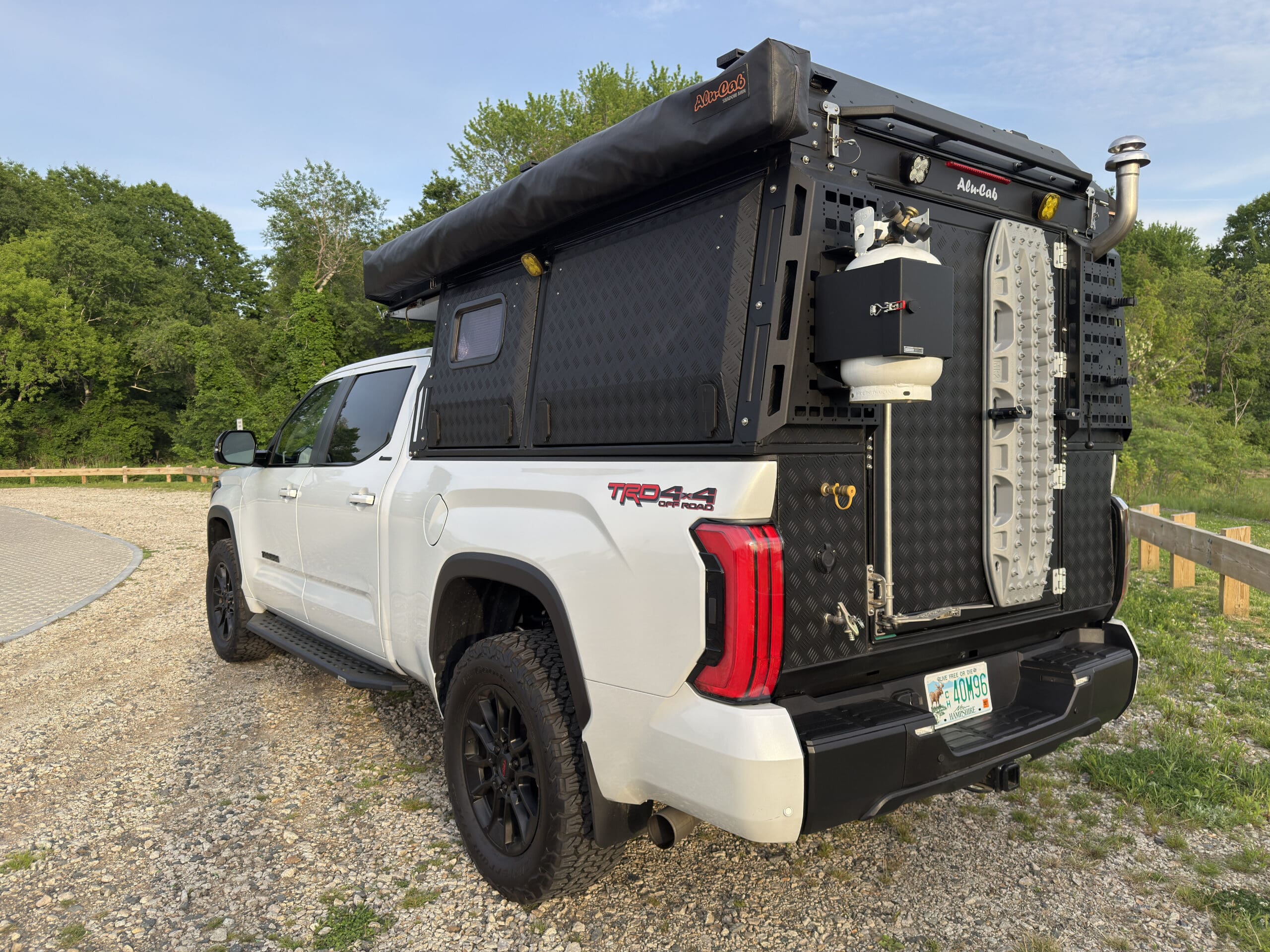 A white Toyota Tacoma TRD 4x4 truck with an Alu-Cab off-road camper setup, outdoor gear attached, and a rooftop tent, parked on a gravel lot near trees and a grassy area.