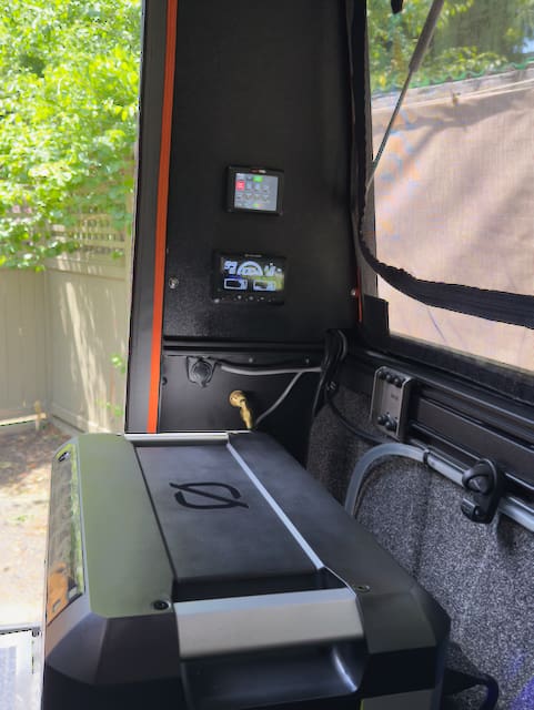 A compact interior space inside the Alu-Cabin with a black and gray cooler or fridge, wall-mounted digital control panels, exposed wiring, and a window revealing greenery and a wooden fence outside.