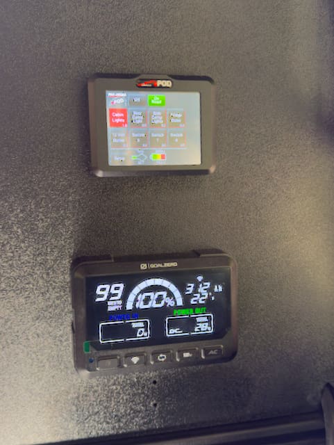 Two electronic control panels mounted on a textured black wall inside the Alu-Cab Alu-Cabin for the 2024 Toyota Tundra. The upper touchscreen displays icons and numbers, while the lower panel shows power and battery info with digital gauges and buttons.