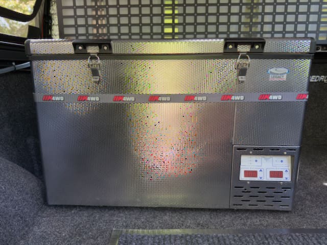 A metallic portable refrigerator with a digital control panel and locks on top, placed in the rear cargo area of a 2024 Toyota Tundra Alu-Cab Alu-Cabin, with a mesh divider behind it. Featured Classified.