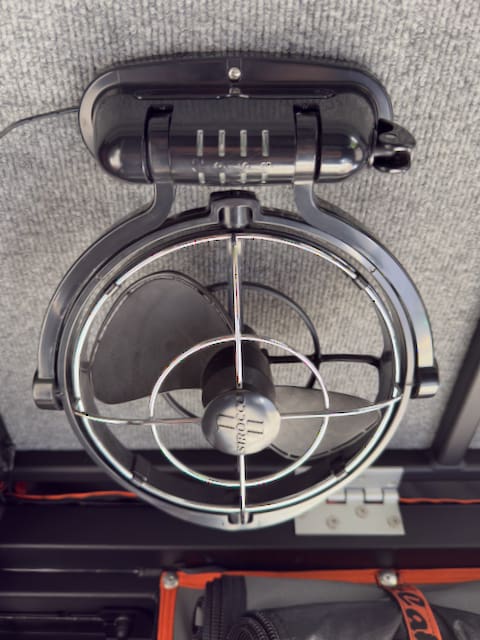 A close-up view of a black, wall-mounted electric fan attached to a gray Alu-Cabin surface, with a protective grill and visible fan blades. Some wiring and an orange-edged bag are partially visible below.