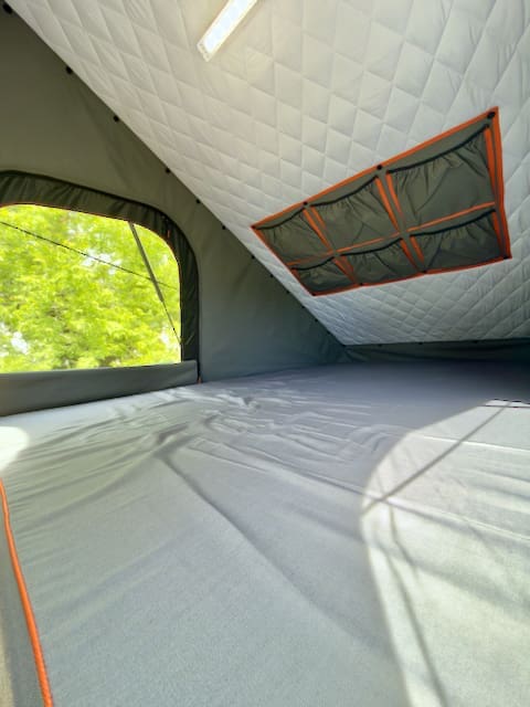 Interior view of a spacious Alu-Cab rooftop tent with quilted white ceiling, mesh storage pockets, and a large window revealing green trees. The sleeping area is covered in gray fabric with orange trim, designed to fit perfectly on a Toyota Tundra or Alu-Cabin.