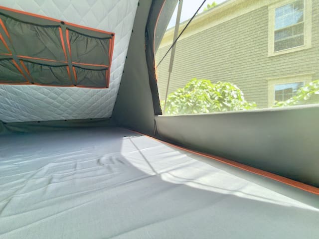 View from inside an Alu-Cab rooftop tent with quilted walls, mesh storage pockets, and a large window revealing sunlight, greenery, and a house with white-framed windows—perfectly set up on your Toyota Tundra.