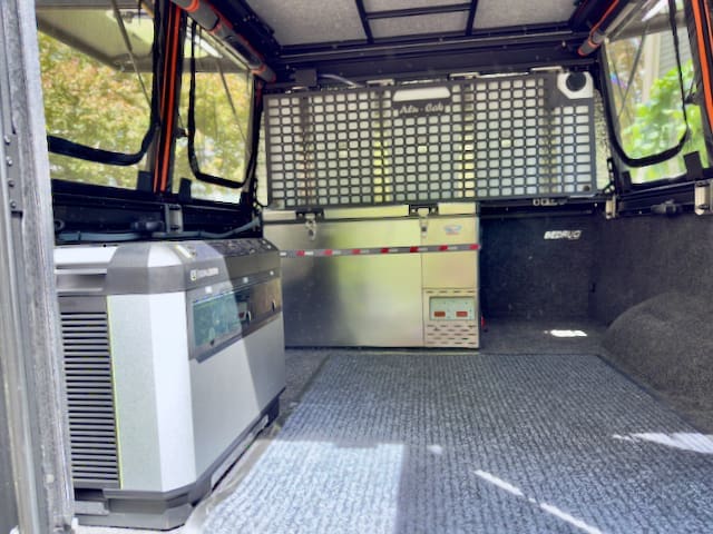 The interior of an Alu-Cab Alu-Cabin’s rear compartment features carpeted flooring, a portable power station on the left, metallic storage at the far end, and mesh panels on the windows and rear divider.