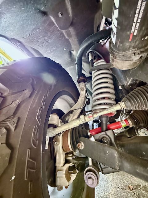 Close-up view of a Toyota Tundra’s front suspension system, showing the coil spring, shock absorber, large off-road tire, control arms, and steering components under the wheel well.