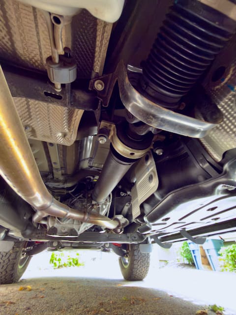View from beneath a Toyota Tundra showcasing the undercarriage, including the exhaust pipe, drive shaft, suspension components, and part of the rear axle. The ground and some greenery are visible below this Alu-Cabin-equipped vehicle.