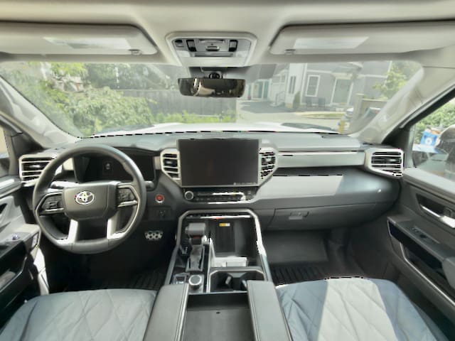 View from the back seat of a modern 2024 Toyota Tundra’s interior, showing the dashboard, steering wheel, large central display, gear shifter, and center console, with houses and greenery visible through the windshield.