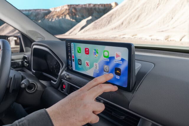 A person’s hand touches a large digital touchscreen display on a car dashboard, showing various app icons and the Overland News feed. Sunlight streams through the window, with barren rocky hills visible outside.