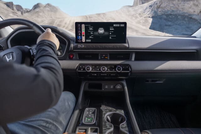 A driver’s view of a modern car interior, featuring a digital dashboard display, climate controls, gear buttons, and a mountainous landscape visible through the windshield—perfect for fans of Overland News.
